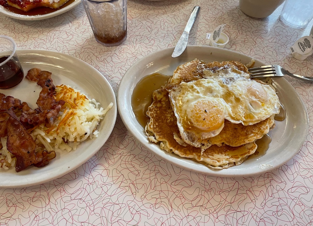 Breakfast with two plates. One plate is pancakes and eggs and the other is hash browns and bacon.